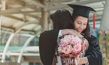 A graduate in a cap and gown hugs another person while holding a bouquet of flowers in an outdoor setting with a covered structure.