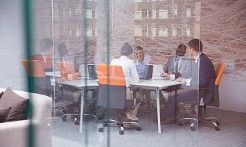 Business professionals discuss and work on laptops around a conference table in a modern office with glass walls and a cityscape view outside.