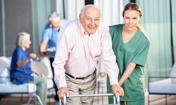 An elderly man uses a walker assisted by a young female caregiver in a green uniform; background shows another elderly couple seated and another caregiver.